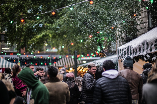 Crowd gathering under colorful string lights at an outdoor market, surrounded by green trees and snow-like effects, while chatting and browsing amidst striped stalls