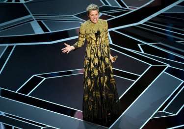 A person in an ornate, long dress stands smiling, gesturing with one hand, holding an award in the other, on a geometrically patterned stage under bright lights.